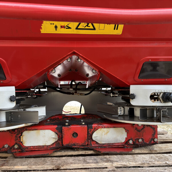 Red industrial machine underside with a yellow safety label and metal brackets visible on a wooden platform.