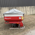 Red Kverneland Exacta-CL agricultural trailer with a gray tarpaulin cover parked on a wooden pallet beside a stone wall and metal building.