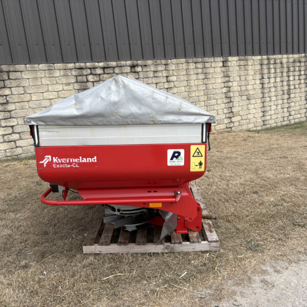 Red Kverneland Exacta-CL agricultural trailer with a gray tarpaulin cover parked on a wooden pallet beside a stone wall and metal building.