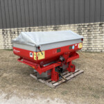 Red agricultural trailer with a grey tarp cover sits on wooden pallets beside a stone wall and dark metal building, outdoors on dry grass.