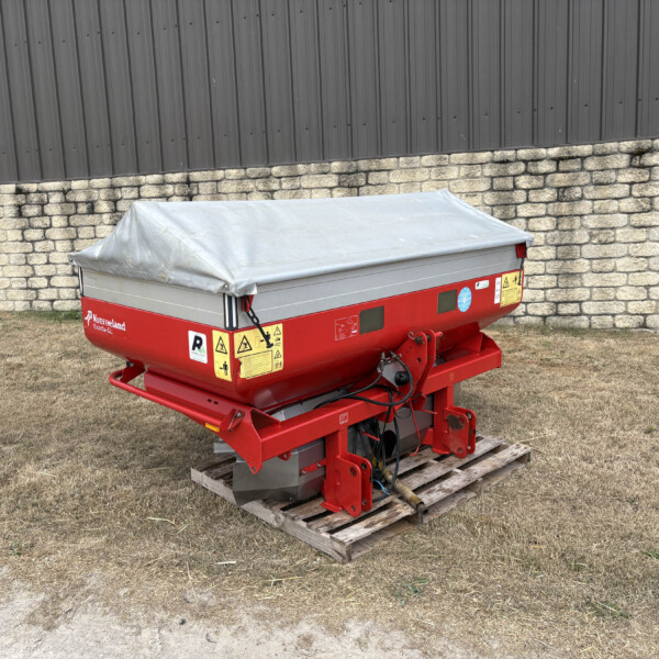 Red agricultural trailer with a grey tarp cover sits on wooden pallets beside a stone wall and dark metal building, outdoors on dry grass.