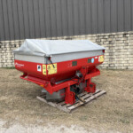 Red farm trailer with a grey tarp cover on a wooden pallet beside a stone wall and dark corrugated metal building in an outdoor lot.