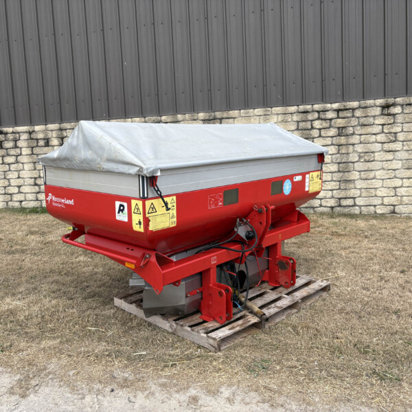Red farm trailer with a grey tarp cover on a wooden pallet beside a stone wall and dark corrugated metal building in an outdoor lot.