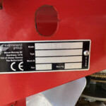 Close-up of a red agricultural machine panel showing a black identification plate reading 'Exacta-CL' with screws and a circular hole nearby, plus an orange reflector on the right.
