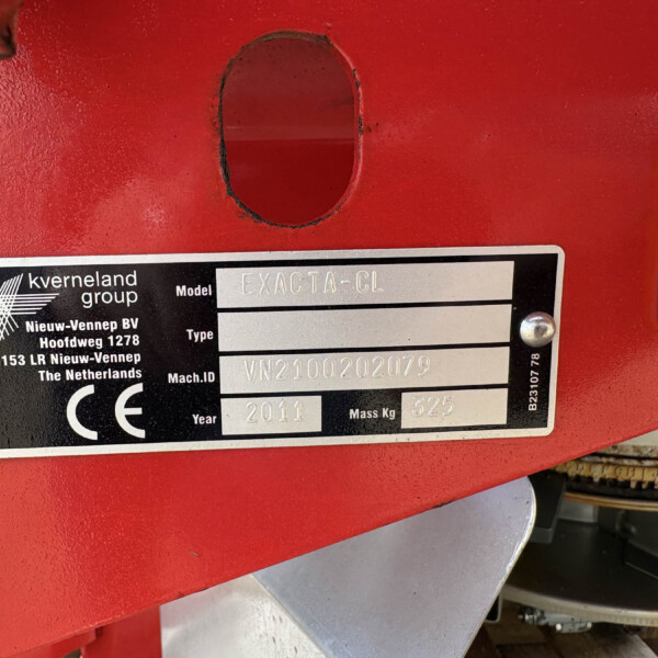 Close-up of a red agricultural machine panel showing a black identification plate reading 'Exacta-CL' with screws and a circular hole nearby, plus an orange reflector on the right.