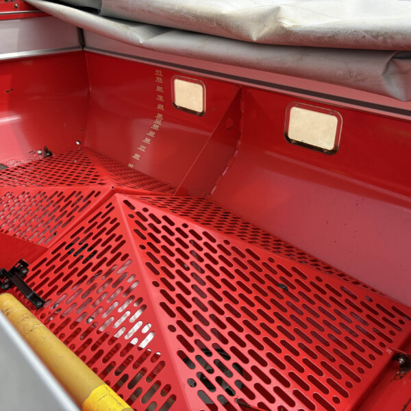 Interior of a red industrial compartment with a perforated red floor, yellow pipe, and a grey, rolled tarp along the top edge.