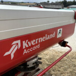 Close-up of a red Kverneland Accord agricultural implement on a trailer, with a rural field and farm buildings in the background.