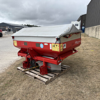 Red agricultural trailer with a grey tarpaulin cover sits on a wooden pallet in a gravel lot beside warehouses and a red utility vehicle in the background.