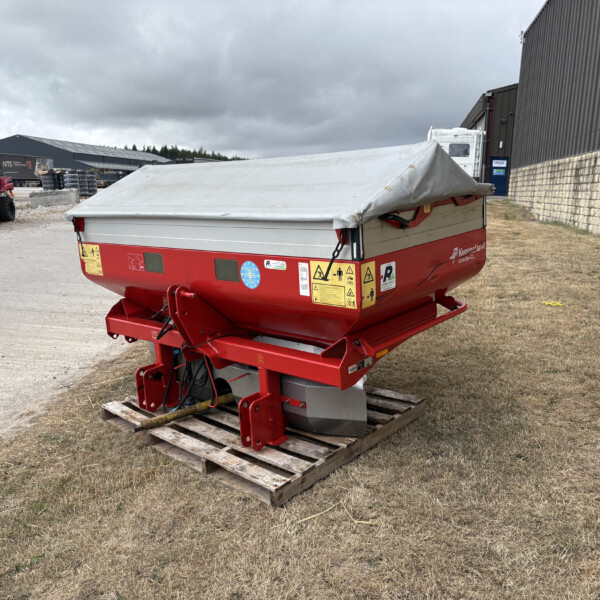 Red agricultural trailer with a grey tarpaulin cover sits on a wooden pallet in a gravel lot beside warehouses and a red utility vehicle in the background.