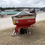 Red Kverneland Exacta-CL farming implement on a wooden pallet in a dusty outdoor yard with green tarps and industrial buildings in the background.