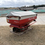 Red Kverneland farming boat on a trailer with a gray cover, placed on a wooden pallet in a dry grassy yard with farm buildings in the background.