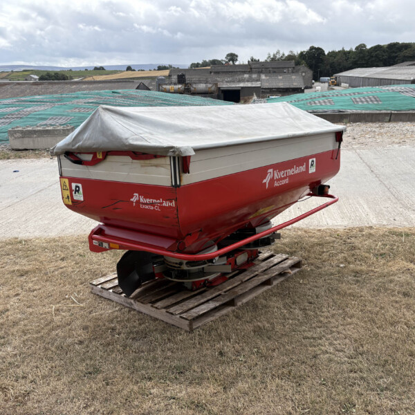 Red Kverneland farming boat on a trailer with a gray cover, placed on a wooden pallet in a dry grassy yard with farm buildings in the background.