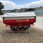 Red Kverneland Accord agricultural implement covered with a white tarp on a wooden pallet in a dusty yard, with a tractor in the background and a large green-surfaced slope.