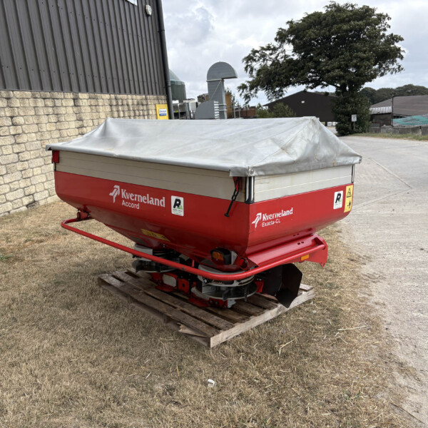 Red Kverneland trailer-mounted agricultural implement with a gray tarp cover, parked on a wooden pallet beside a brick wall in an industrial area.
