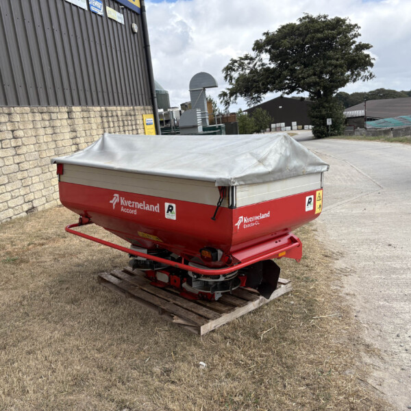 Red Kverneland Exacta-Ci implement with gray tarp cover on a wooden pallet in an outdoor yard near a brick wall and tractor tire edge.