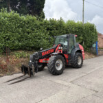 Red Manitou tractor with front loading forks on a street beside a tall hedge and brick buildings in the background.