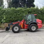 Red Manitou forklift with forks resting on the ground on a paved lot, hedge in the background.