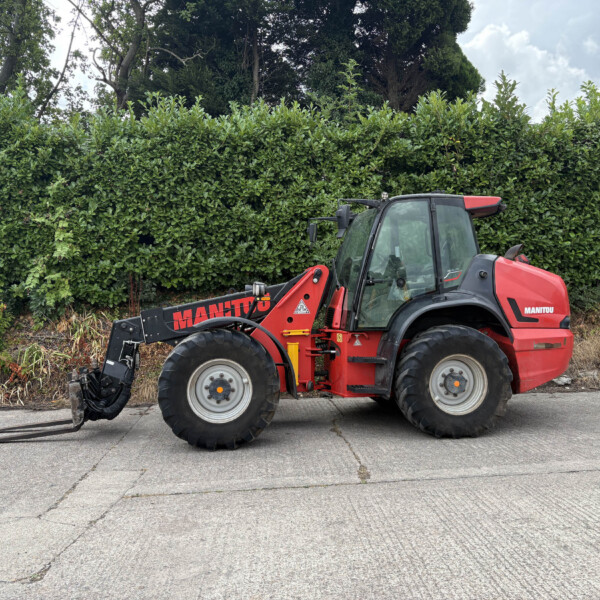 Red Manitou forklift with forks resting on the ground on a paved lot, hedge in the background.
