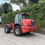 Red Manitou agricultural tractor parked in a lot beside a tall green hedge and a white building with a yellow sign; license plate DU23 DSW visible on the cab front.