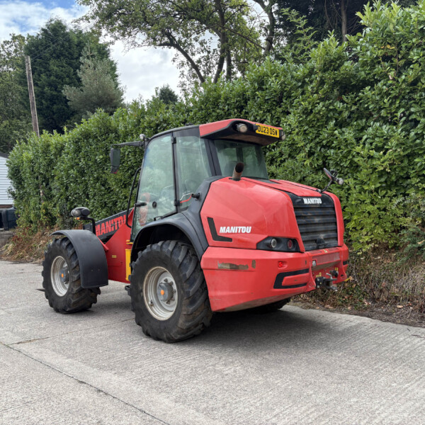 Red Manitou agricultural tractor parked in a lot beside a tall green hedge and a white building with a yellow sign; license plate DU23 DSW visible on the cab front.