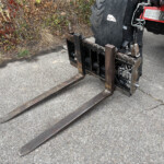Rusty forklift forks extend over a paved surface with a leafy hedge in the background.