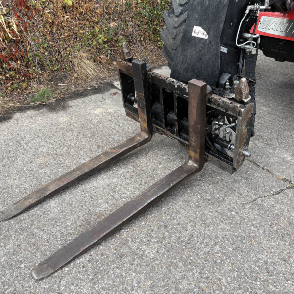 Rusty forklift forks extend over a paved surface with a leafy hedge in the background.