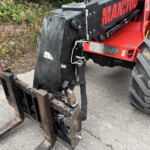Red Manitou telehandler with a large front fork attachment resting on the ground beside overgrown bushes.