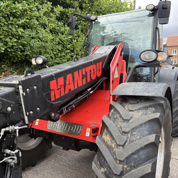 Close-up of a red Manitou telehandler arm attached to a black industrial vehicle, with green hedge and brick building in the background.