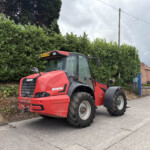 Red Manitou telehandler parked along a street beside tall green hedge under a cloudy sky, with brick buildings in the background.