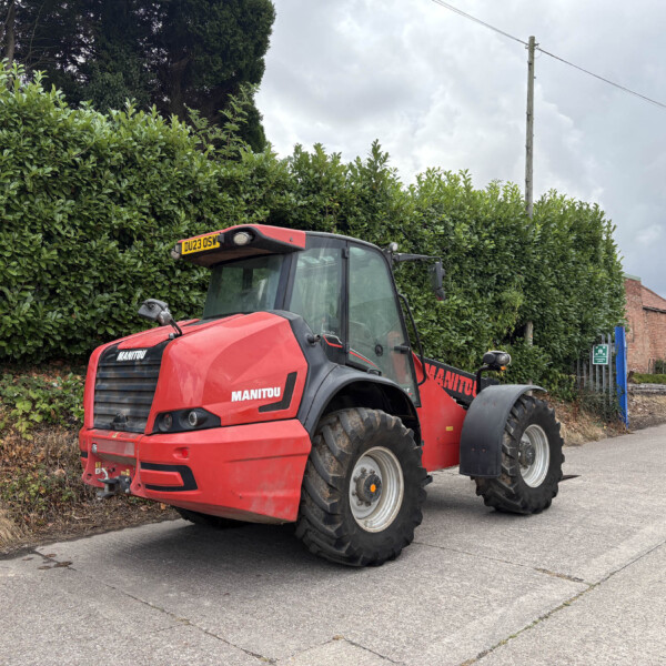 Red Manitou telehandler parked along a street beside tall green hedge under a cloudy sky, with brick buildings in the background.
