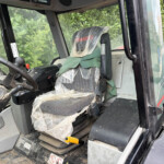 Cabin interior of a large construction machine with a plastic-wrapped seat, dusty controls, and a worn steering wheel overlooking a dusty floor area.