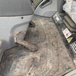 Mud-splattered interior of a construction vehicle cabin showing a dirty floor mat, seat, and yellow control knob with warning stickers nearby.