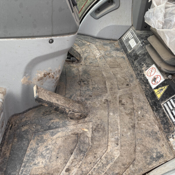 Mud-splattered interior of a construction vehicle cabin showing a dirty floor mat, seat, and yellow control knob with warning stickers nearby.