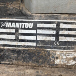 Brand label on a muddy machine showing 'MANITOU' with specifications (unladen mass ~8430 kg, power ~105 kW) and serial/model information partially obscured by dirt.