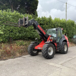 Red Manitou telehandler with extended pallet forks raised beside a dense green hedge along a street.
