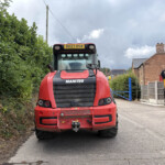 Front view of a red Manitou telehandler parked in a residential driveway, with a tall hedge on the left and brick houses to the right under a cloudy sky.