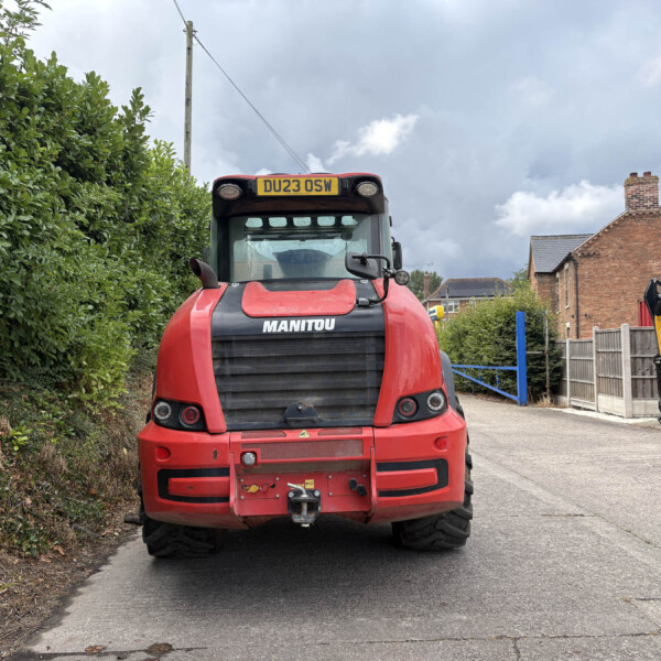 Front view of a red Manitou telehandler parked in a residential driveway, with a tall hedge on the left and brick houses to the right under a cloudy sky.