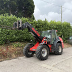 Red telescopic loader with extended forks raised beside a tall evergreen hedge in a construction area; cloudy sky in the background and a brick building to the right.
