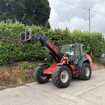 Red telescopic loader with extended forks raised beside a tall evergreen hedge in a construction area; cloudy sky in the background and a brick building to the right.