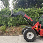 Red Manitou brand construction loader with extended fork attachment beside a green hedge, parked on a paved area.