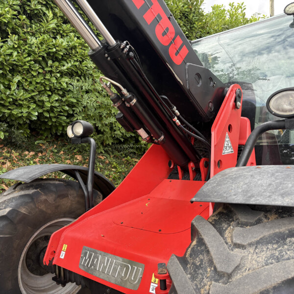 Red Manitou telescopic handler with hydraulic arm resting beside a green hedge, showing cabin and large tires in a construction setting.