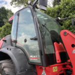 Close-up of a red tractor cab with large curved windshield, wipers, and a front loader arm against a green hedge.