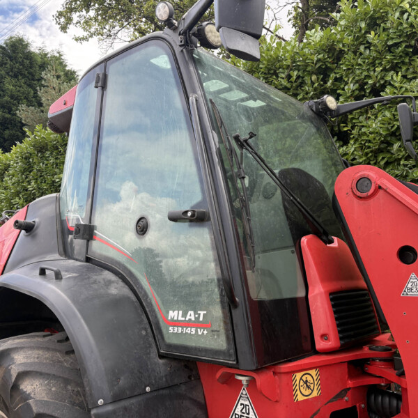 Close-up of a red tractor cab with large curved windshield, wipers, and a front loader arm against a green hedge.