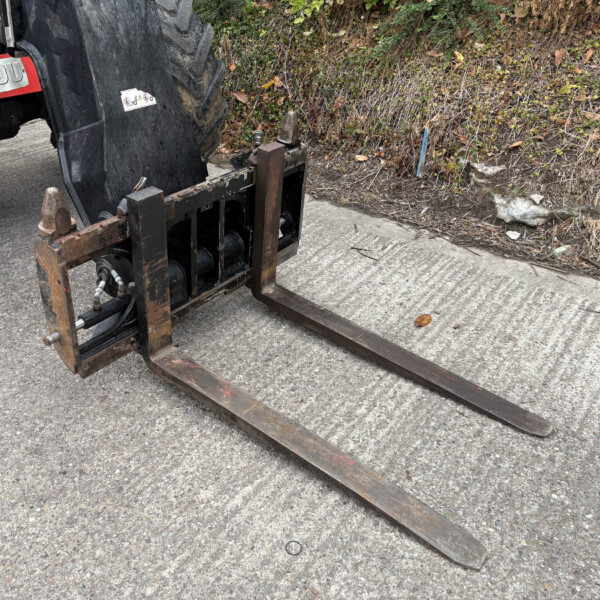 Forklift pallet forks resting on a rough concrete surface beside a hedge, with a red Manitou machine visible in the background.
