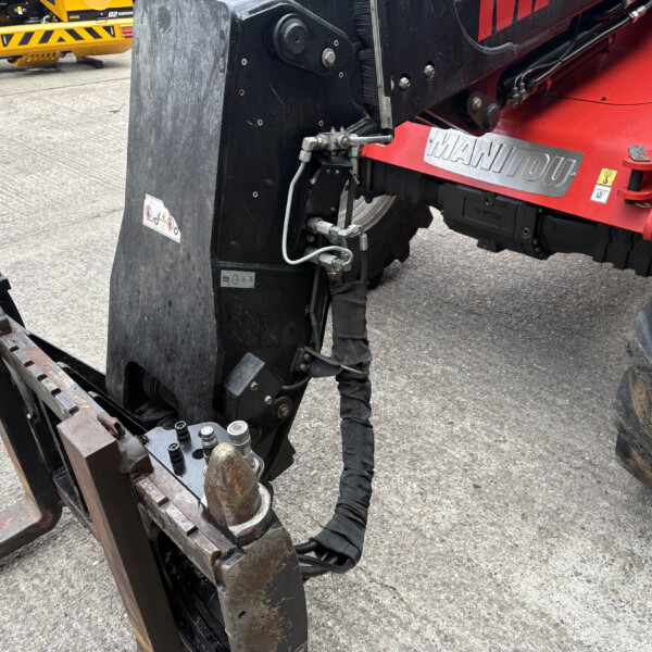 Close-up of a red Manitou telehandler with a rusted bucket attachment and visible hydraulic hoses on a paved surface, a worker's leg nearby.