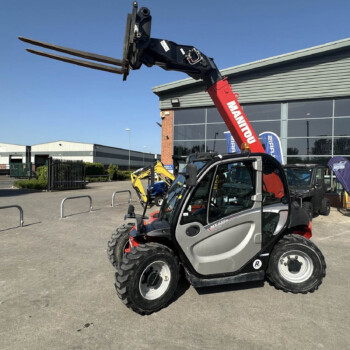 Red forklift arm with a three-pronged fork attachment raised over a compact vehicle in a sunny outdoor industrial lot near a modern building.