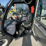 Cab interior of a large construction machine with steering wheel and controls; visible yellow excavator outside through the window