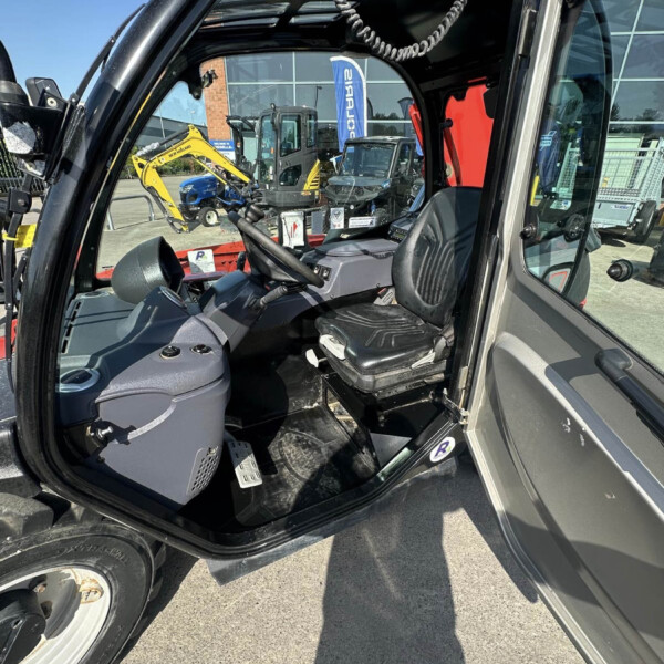 Cab interior of a large construction machine with steering wheel and controls; visible yellow excavator outside through the window