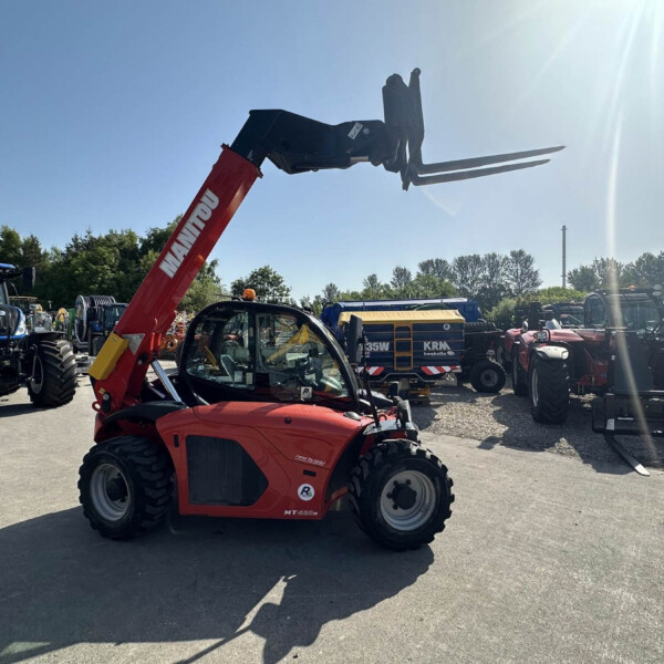 Red Manitou telescopic forklift with extended forks in a sunny outdoor machinery yard