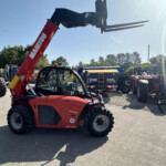 Red Manitou telescopic handler with forks raised, in a yard full of tractors and farm machinery under a clear blue sky.
